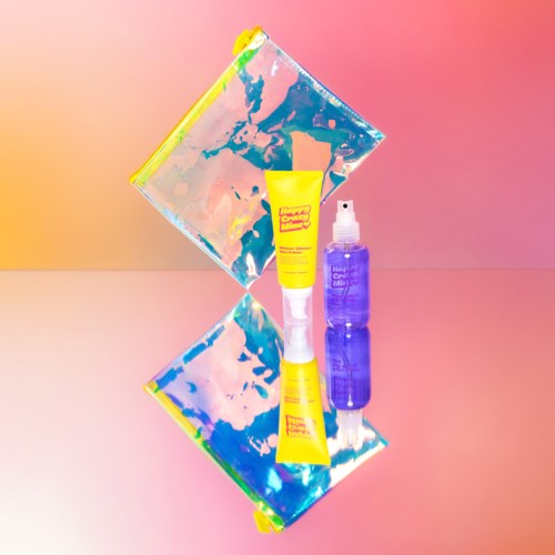 Een gele tube en een paarse sprayfles liggen op een spiegel met kleurrijke, abstracte achtergrond en reflectie.