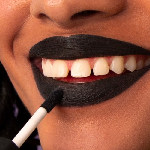 A person applies black lipstick on their lips, showing teeth with some gaps and skin around the mouth.