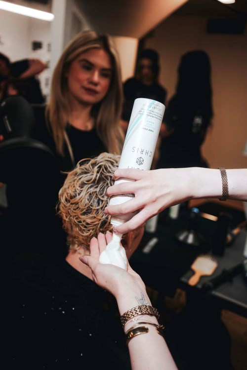 A person is applying hair spray to a curly-haired individual in a busy salon. A woman watches nearby.