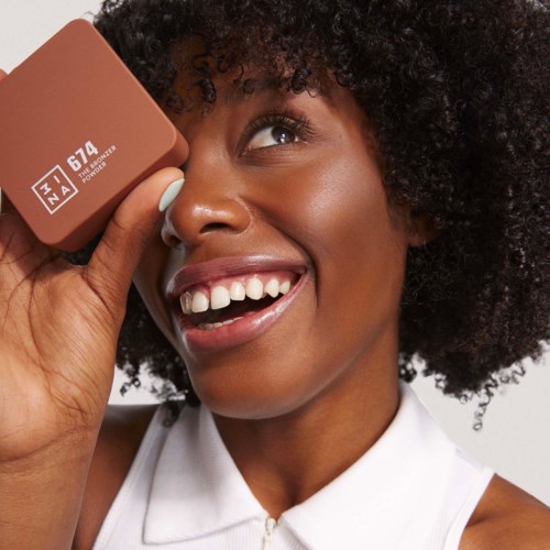 En smilende kvinne med krøllete hår holder et makeup-produkt nær øyet, viser hvite tenner og glede.