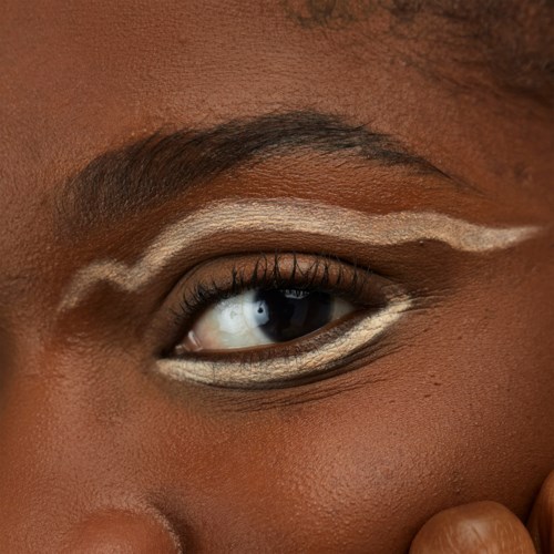 A close-up of a person's eye with white face paint or makeup lining the eye and eyebrow, and a brown skin tone.