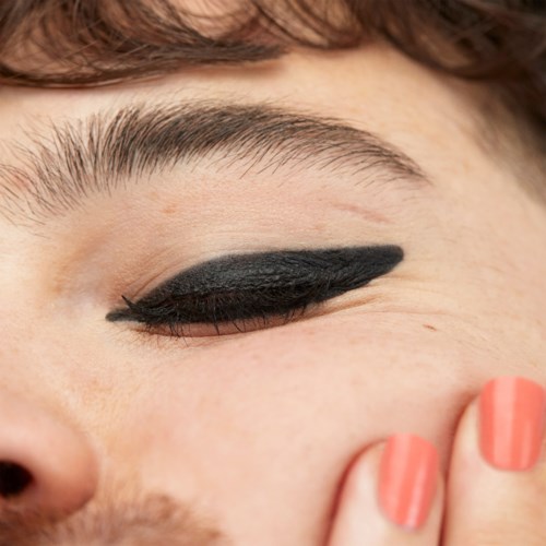 Close-up of a person with black eyeliner, showing their eye and part of their face with a hand.