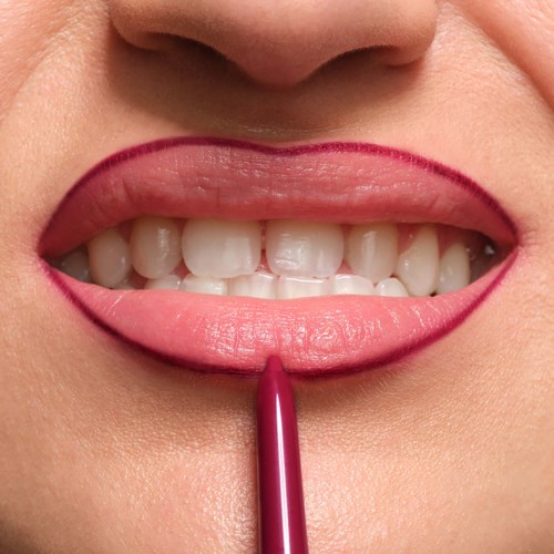 A person is smiling with lipstick and lip liner, showing teeth, with a lip liner pencil touching the lips.