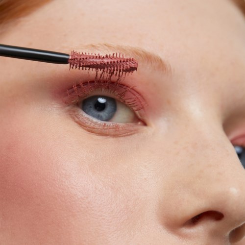 A person is applying pink mascara to their upper eyelashes with a brush.