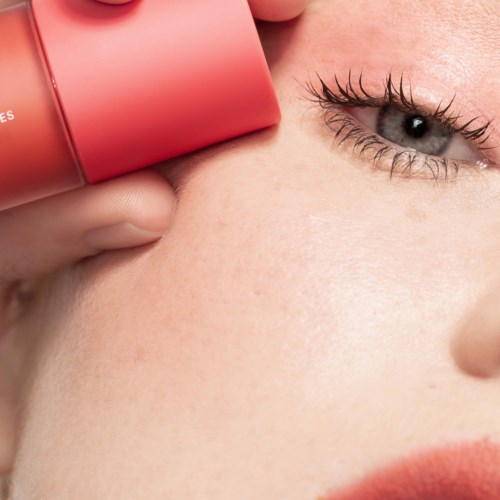 A person applying a pink skincare product near one eye, close-up of the face with clear skin and makeup.