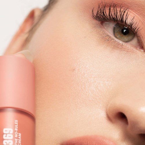 Närbild av en kvinnas ansikte med makeup, med fokus på ögat och huden, som håller en rosa hudvårdsprodukt.