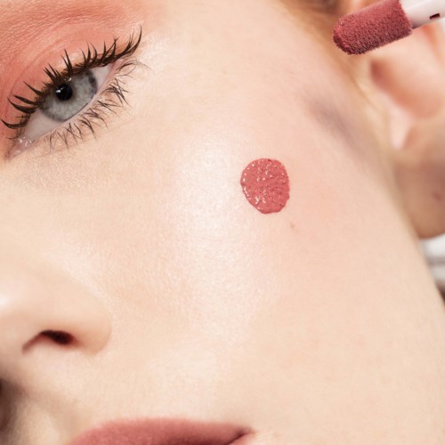 A person is applying blush on the cheek, near a red beauty mark on their face.