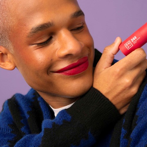 A person with lipstick applies a red lipstick on their lips, smiling happily. They wear a black and blue sweater.