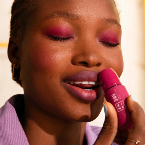 A woman with pink eye makeup and gold earrings holds a pink lipstick close to her lips.