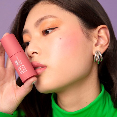 A woman with makeup holding a pink spray can, showing her face and ear against a purple background.
