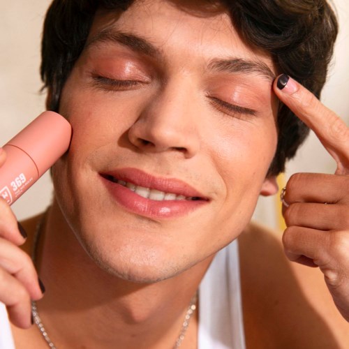 A person with short dark hair smiling, holding a lipstick, and touching their temple.