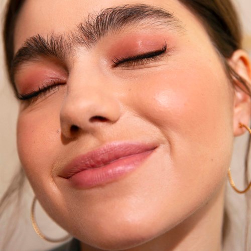 Close-up of a woman's face with closed eyes, showing clear skin, soft makeup, and gold hoop earrings.