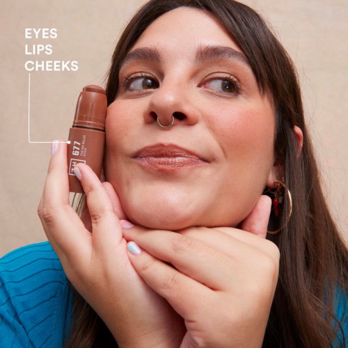 A woman holds a lipstick near her face, with labels pointing to her eyes, lips, and cheeks, smiling gently.
