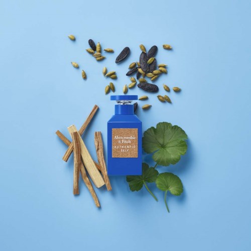 Een blauwe fles met label "Abercrombie & Fitch", omringd door houten stokjes, zaden en groene bladeren op een lichtblauwe achtergrond.