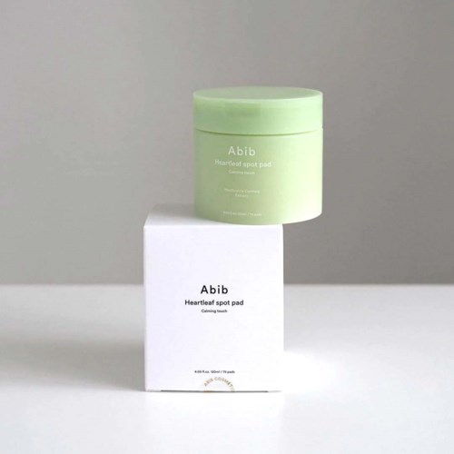 Een groene pot en een wit doosje, allebei gelabeld Abib Heartleaf spot pad, staan tegen een neutrale achtergrond.