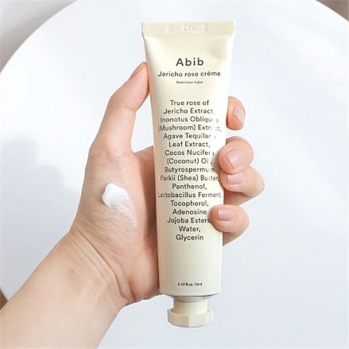 Een hand houdt een tube Abib Jericho Rose Crème, met wat crème op de vingers en een witte achtergrond.