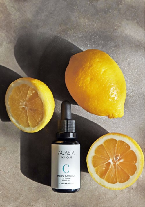 Eine Zitrone, halbierte Zitrone und Zitronenscheibe umgeben eine Hautpflege-Serum-Flasche auf einer grauen Oberfläche.