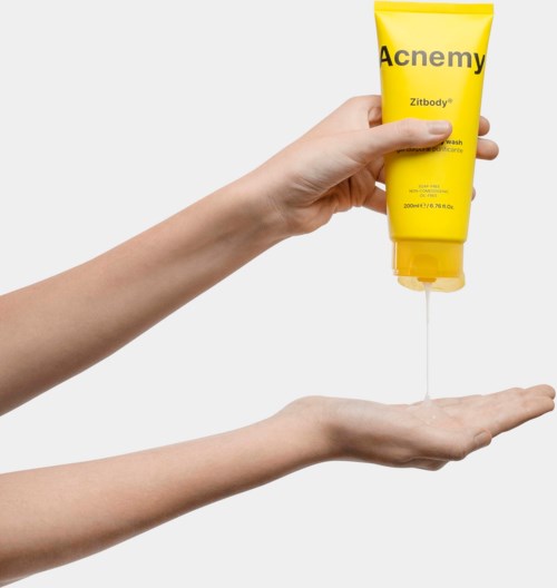 Iemand knijpt een gele tube met 'Acne'my' erin in de hand. De vloeistof komt uit de tube.