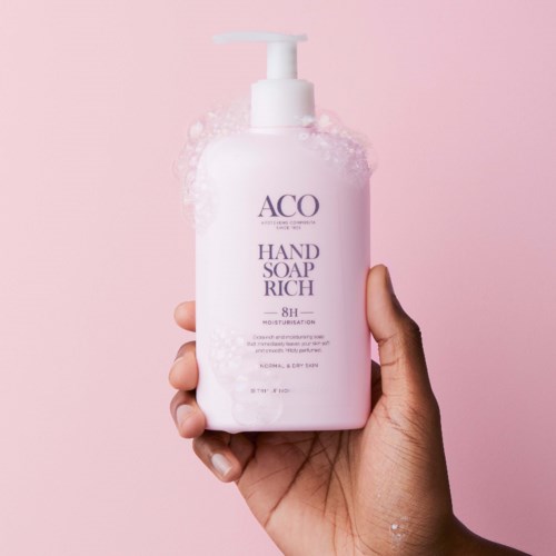 En hand håller en rosa flaska märkt 'ACQ Hand Soap Rich' med pump mot en rosa bakgrund.