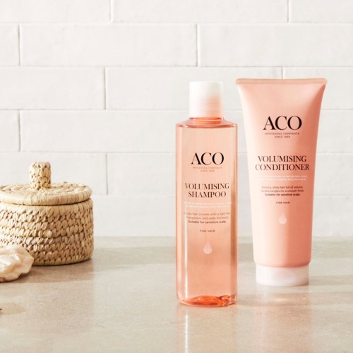 Two pink hair care products, shampoo and conditioner, are on a light surface with a woven basket and stones nearby.