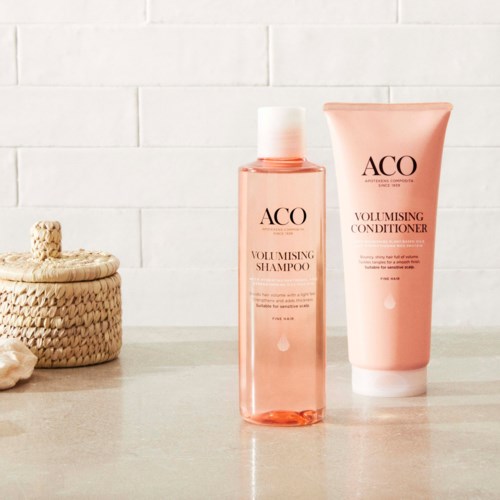 Two pink hair care bottles on a light countertop with a woven basket and white brick wall behind.