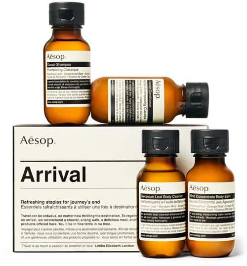 Vier braune Flaschen mit Aesop-Produkten auf einem weißen Ständer, einschließlich Shampoo, Körperreiniger und Körperbalsam.