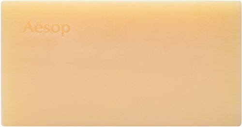 Dies ist ein Logo mit dem Wort "Aesop" in Orange auf einem hellen, pfirsichfarbenen Hintergrund.