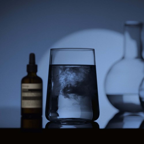 Ein Wasserglas mit einem trüben Wirbel innen, auf einer dunklen Oberfläche, dahinter eine braune Tropfflasche und daneben eine runde Flasche.