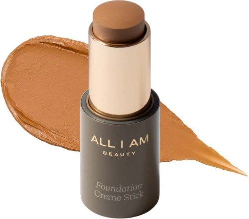 A foundation stick with brown color, labeled 'ALL I AM BEAUTY,' placed in front of a smear of matching foundation.
