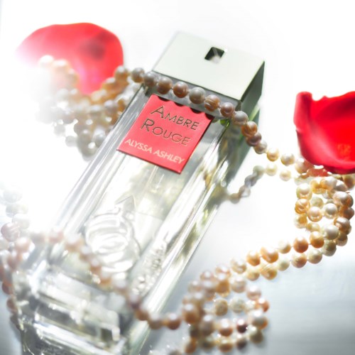 A clear perfume bottle labeled 'Amber Rouge' by Alyssa Ashley, surrounded by pearl necklaces and rose petals.