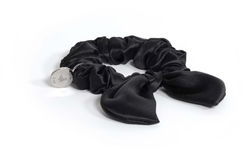 En svart satinscrunchie med rosett, fäst vid en liten silverhänge, som ligger på en vit yta.