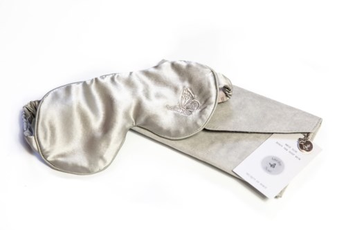 En glänsande silver sömnmask bredvid ett grått fodral och ett kort. Masken har en broderad logga eller bild.