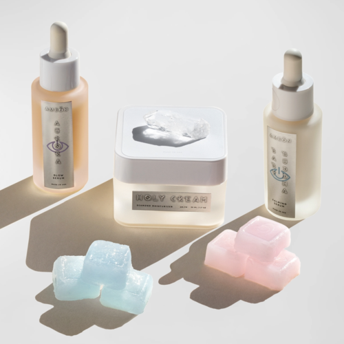 Zwei Hautpflegeflaschen, ein Glas mit 'Holy Cream' und bunte Seifen in Blau und Rosa. Schlichte und saubere Anordnung.