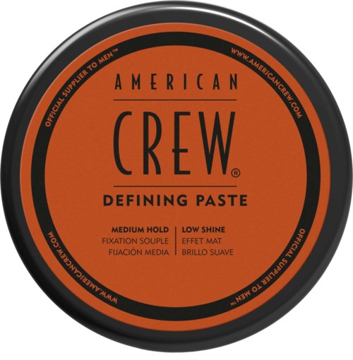A round container lid with black and orange colors, labeled 'American Crew Defining Paste' for hair styling.