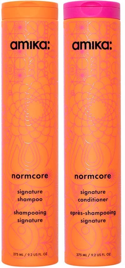 Kaksi oranssia Amika Normcore shampoopulloa ja hoitoainetta, pinkit kanteet.