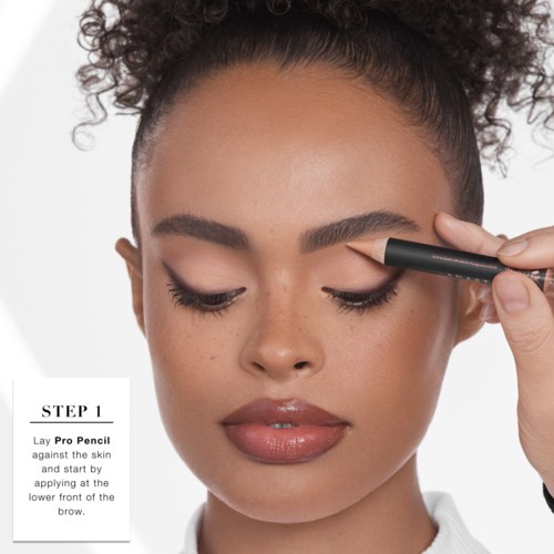 Eine Frau zeichnet Augenbrauen mit einem schwarzen Bleistift, ihre Augen sind geschlossen, zeigt Make-up und lockiges Haar.