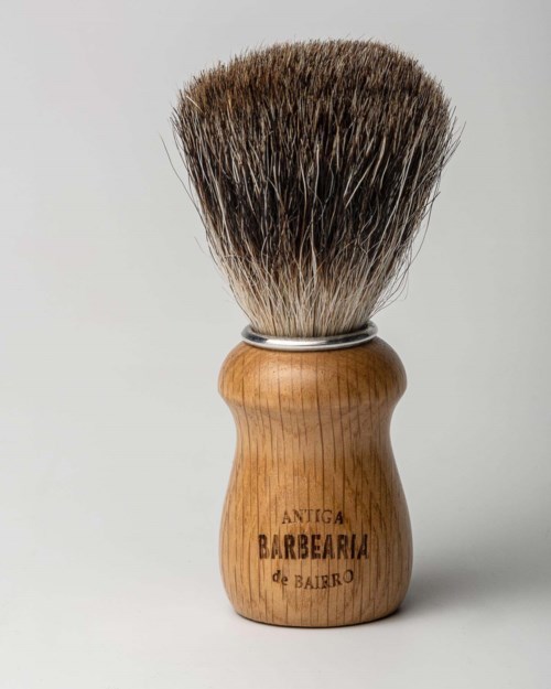 A shaving brush with a wooden handle and dense bristles. The handle has the words "Antiga Barbearia de Bairro" engraved.