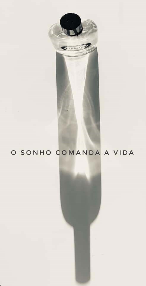 A clear glass bottle with a black cap casts a shadow resembling a dagger. The text says "O SONHO COMANDA A VIDA."