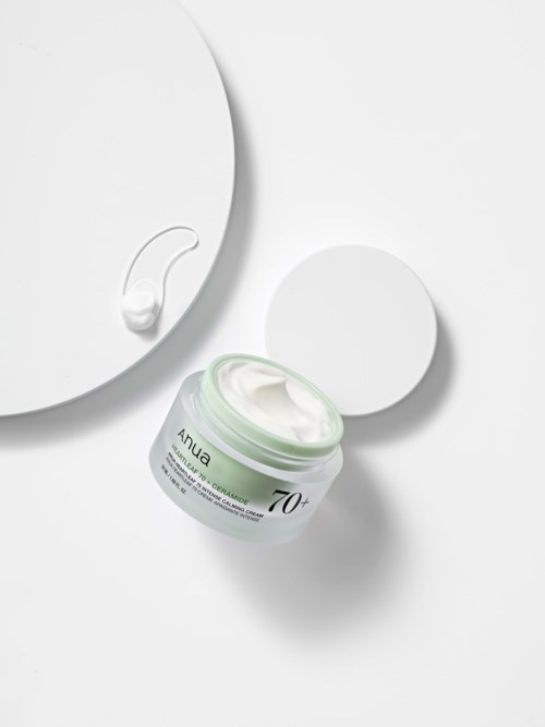 Een potje met crème met label 'Anua 70+', met een beetje crème op een lepel en een cirkelvormig object in de buurt.