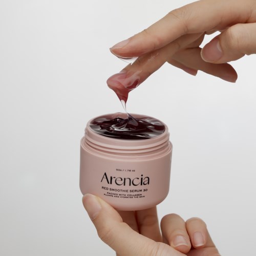 Eine Hand taucht den Finger in einen pinken Behälter mit dunkelroher Gel oder Creme für Hautpflege. Der Behälter ist mit Arencia beschriftet.