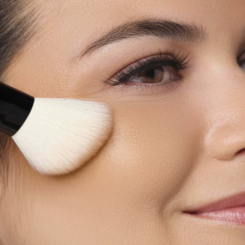 Een vrouw brengt make-up aan met een kwast op haar wang, glimlachend zacht met een glad, heldere huid en verzorgde wenkbrauwen.