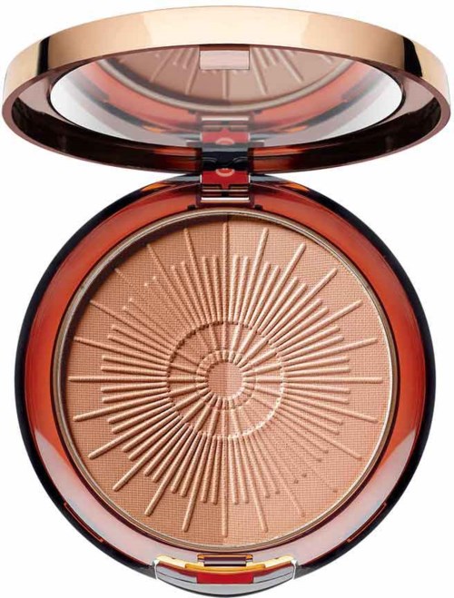 A compact powder with a decorative sunburst pattern inside, in a shiny, round case. The lid is open, showing the product.