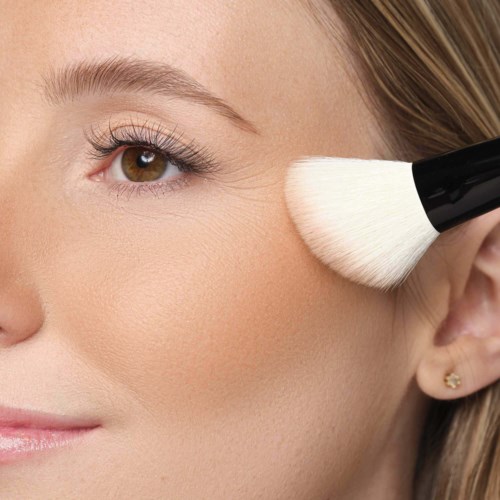 A woman uses a brush to apply makeup under her eye, with focused attention on her makeup routine.