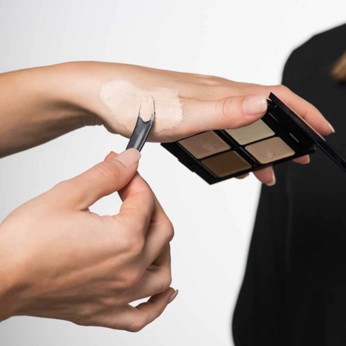 Någon applicerar foundation eller concealer på armen med en liten borste från en ögonskuggspalett.