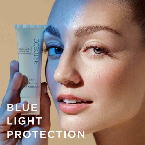 A woman holds a skincare tube labeled 'Blue Light Protection,' smiling slightly, with clear skin and bright lighting.