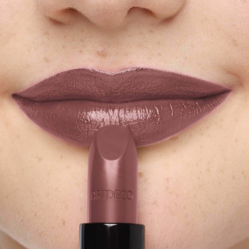 Iemand brengt bruin lippenstift aan op de lippen, die vol en licht geopend zijn. De verpakking van de lippenstift is zichtbaar.