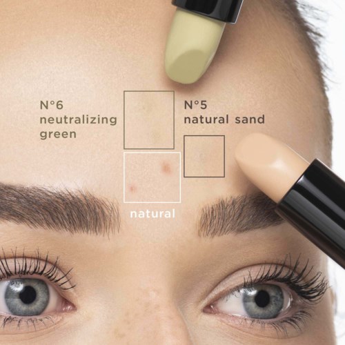Das Bild zeigt die Stirn einer Person, die mit verschiedenen Make-up- oder Concealer-Nuancen getestet wird. Textetiketten zeigen die Farbtöne. Zwei Makeup-Stifte sind auf die Stirn aufgetragen.