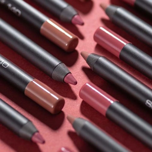 Meerdere lippenpotloodjes in verschillende tinten gerangschikt op een roze achtergrond.