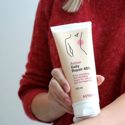 A person in a red sweater holds a tube of skin repair cream with a minimalist line drawing on it.