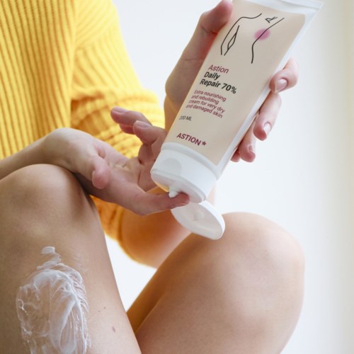 Someone applying cream to their legs, holding a white tube with text and a diagram on it.